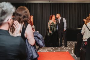 Bride in a black floral gown laughing with the groom at the end of the ceremony aisle as guests applaud