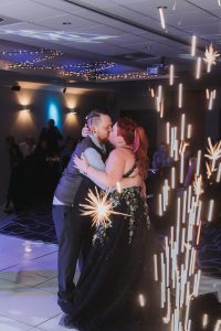 Bride in a black floral gown kissing the groom amid a cold-sparkler shower on the dance floor