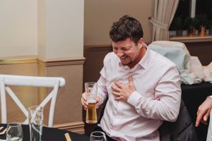 Wedding guest in a pink shirt laughing as he holds a pint of beer at the reception