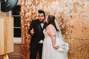 Photo-booth portrait of the bride and groom at a gold sequin backdrop with a 'Mr & Mrs' neon sign