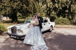 Bride and groom posing in front of a vintage white Rolls-Royce wedding car under a tree