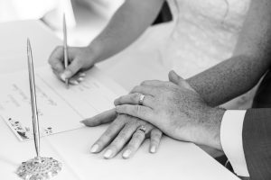 Black and white close-up of the bride signing the marriage register with the groom's hand on hers