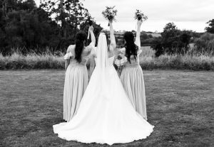 Black and white photo of the bride with two bridesmaids raising bouquets in the countryside