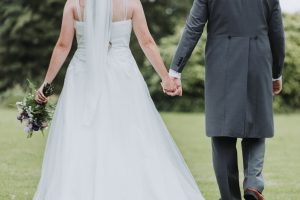Bride with bouquet and groom in a tailcoat walking hand in hand on a lawn, viewed from behind