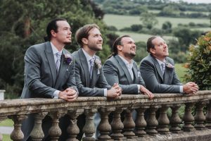 Groomsmen in grey morning suits and lilac ties leaning on a stone balustrade overlooking countryside