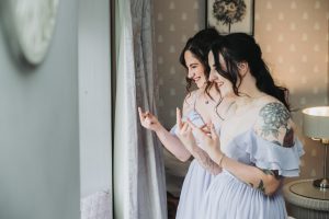 Two bridesmaids in lilac dresses laughing while peeking through a window curtain