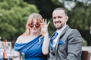 Wedding guests posing playfully — woman in a blue dress lowering sunglasses beside a man in a grey suit
