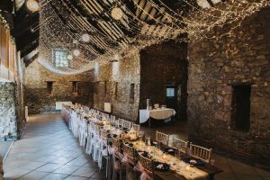 Wide view of a stone barn wedding venue with a fairy-light ceiling and a long banquet table
