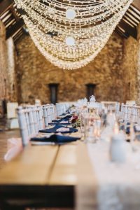Long banquet table set for a wedding reception in a rustic stone barn under a twinkle-light canopy