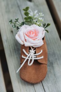 Wedding detail of a baby's brown suede shoe with a pink rose accent on weathered wood