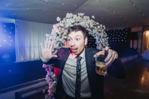 Groom posing with a whiskey glass under a floral arch at an evening wedding reception